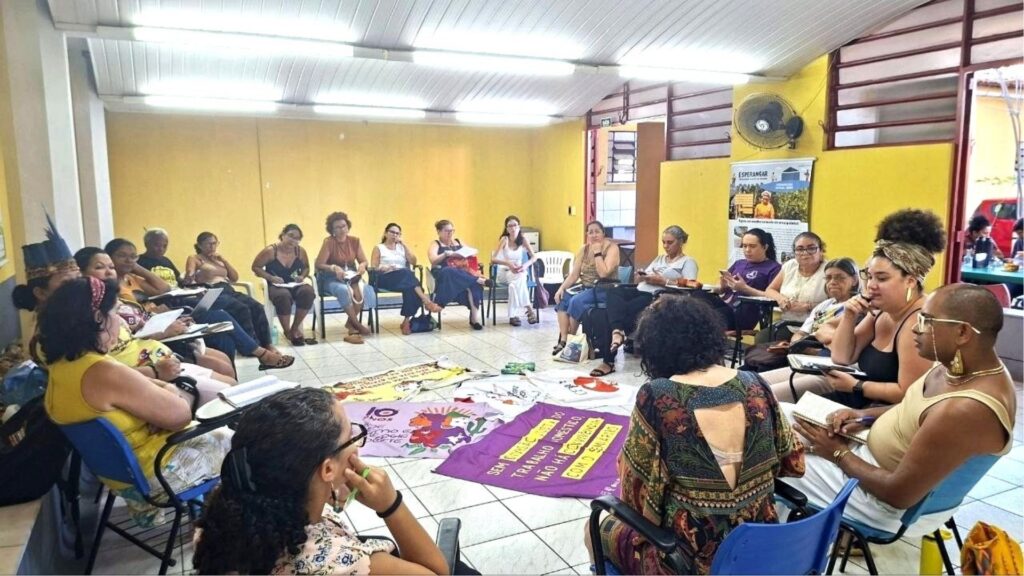Grupo de mulheres sentadas em círculo dentro de uma sala com paredes amarelas, participando de uma oficina ou roda de conversa. Algumas seguram cadernos e canetas, enquanto faixas com mensagens como “Sem trabalho decente não há conquistas” estão estendidas no chão ao centro do grupo.