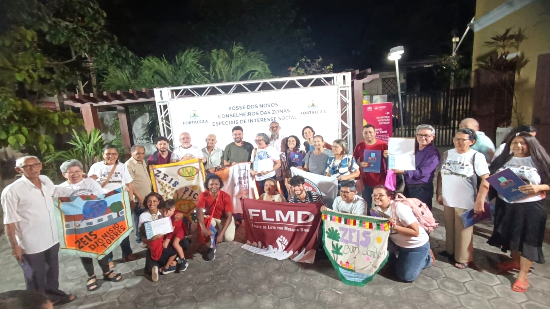Grupo diverso de pessoas posando ao ar livre à noite, segurando faixas com os nomes de movimentos sociais e banners da Prefeitura de Fortaleza. Ao fundo, um painel anuncia a "Posse dos Novos Conselheiros das Zonas Especiais de Interesse Social".