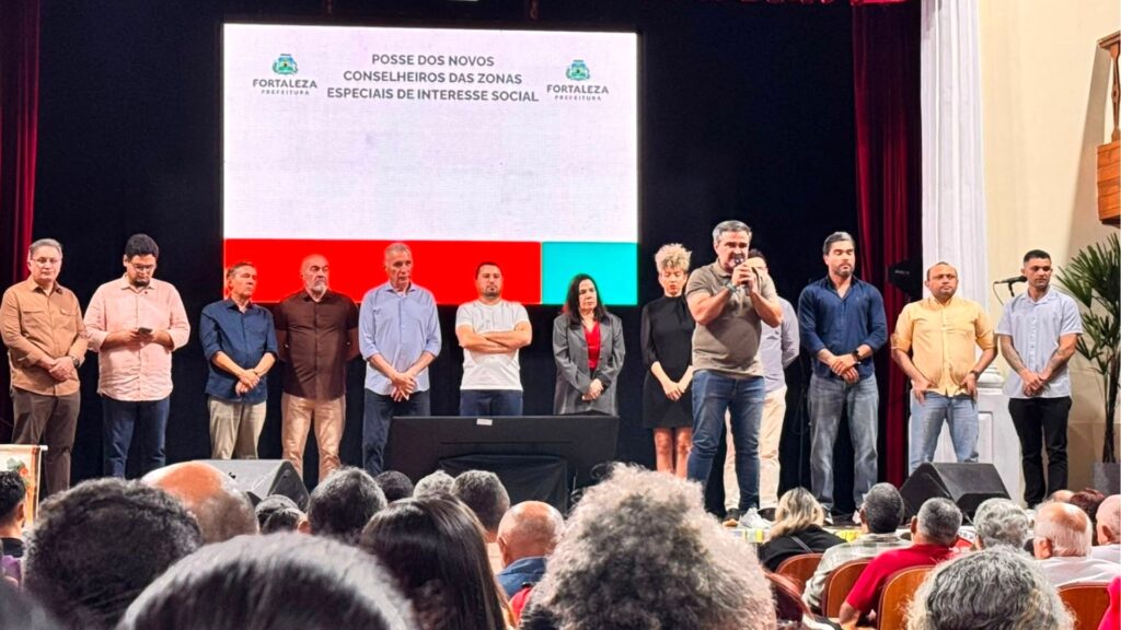 Homens e mulheres em pé sobre um palco durante cerimônia oficial, com um grande telão ao fundo exibindo o título "Posse dos Novos Conselheiros das Zonas Especiais de Interesse Social" e o logo da Prefeitura de Fortaleza. Um homem no centro segura um microfone enquanto fala para o público sentado nas fileiras abaixo.