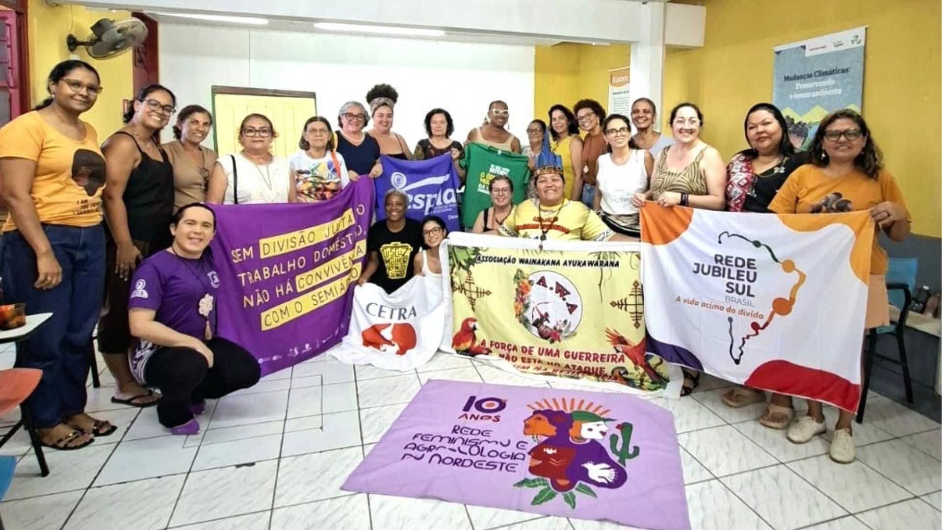 Grupo de mulheres posando para foto dentro de uma sala com paredes amarelas, segurando diversas faixas e bandeiras de organizações sociais. Entre as faixas visíveis estão a da "Rede Jubileu Sul Brasil", com o lema "A vida acima da dívida", outra roxa com a frase "Sem divisão justa do trabalho doméstico não há convivência sem violência", além de banners de associações indígenas e feministas estendidos no chão ao centro do grupo.