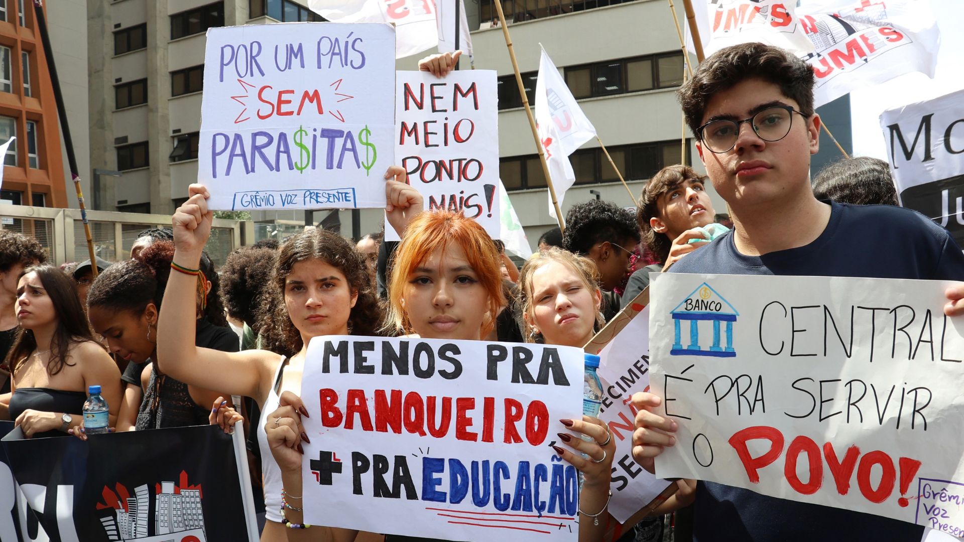 Grupo de jovens manifestantes segurando cartazes com mensagens políticas e econômicas. Uma jovem no centro segura um cartaz que diz "Menos pra banqueiro + pra educação", enquanto um rapaz à direita exibe outro com a frase "Banco Central é pra servir o povo!". Ao fundo, outros participantes carregam faixas e bandeiras em uma rua urbana. Foto: Rovena Rosa/Agência Brasil