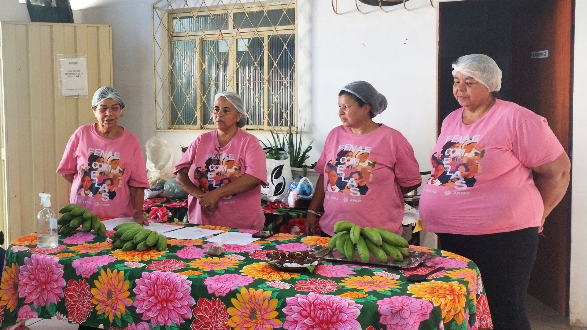 Quatro mulheres usando camisetas rosa com a inscrição "FENAE COM ELAS" e toucas de cabelo estão atrás de uma mesa coberta por um pano floral. Sobre a mesa, há cachos de bananas verdes, pães e outros alimentos, sugerindo uma oficina ou evento comunitário. Ao fundo, vê-se uma janela com grades e uma porta.